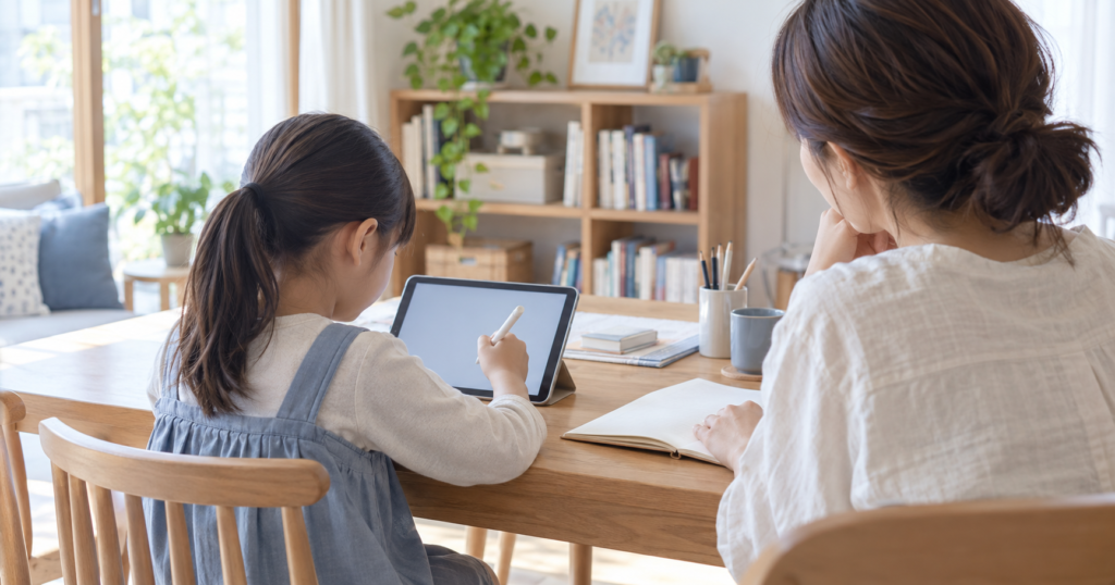 自宅のテーブルでタブレットに取り組む小学生と、隣で見守る保護者