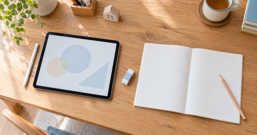 木の机に並べたタブレットとタッチペン、方眼ノートと鉛筆